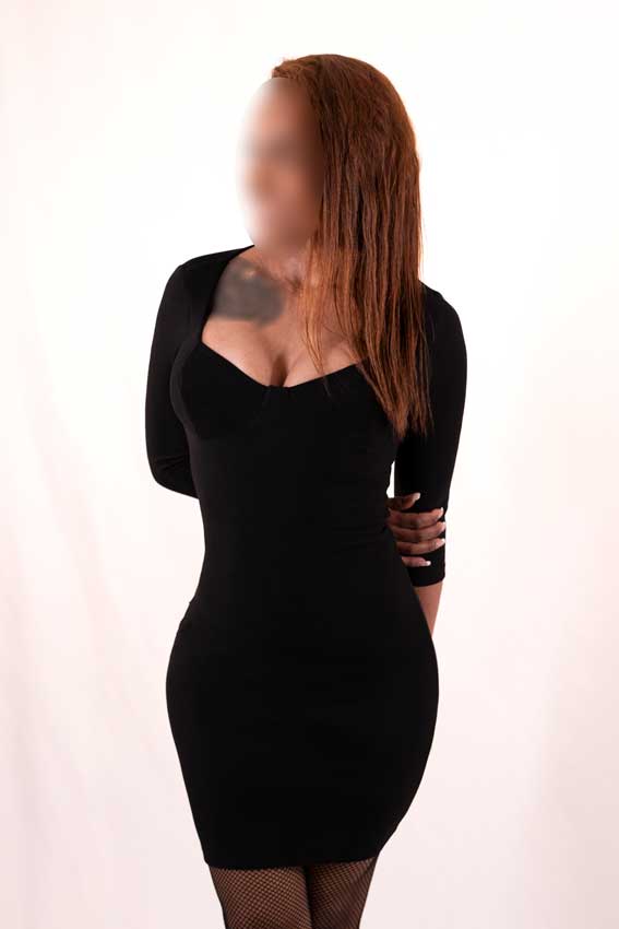Alicia, escort med brunt og bølget hår poserer siddende på en sort stol i Málaga